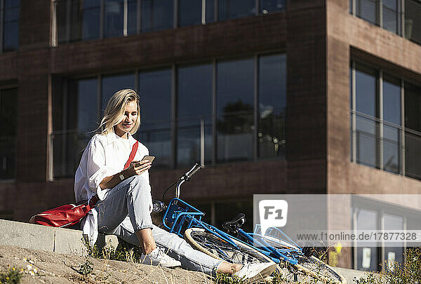 Smiling blond woman using smart phone sitting by bicycle