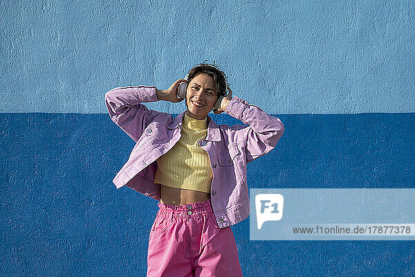 Smiling woman listening music through headphones in front of blue wall