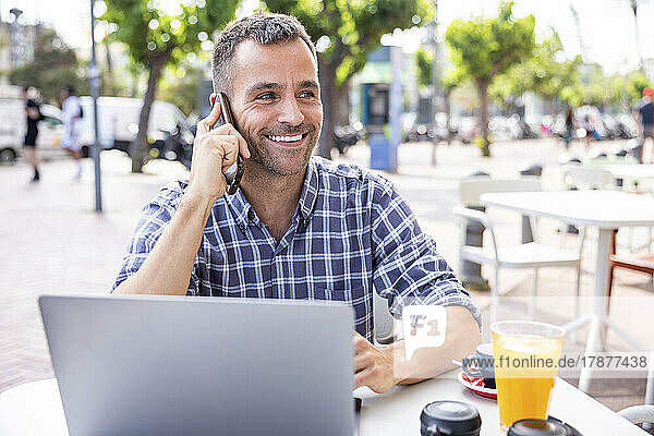 Smiling mature freelancer talking through smart phone sitting with laptop at outdoor cafe