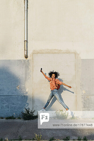 Happy woman holding smart phone jumping in front of wall