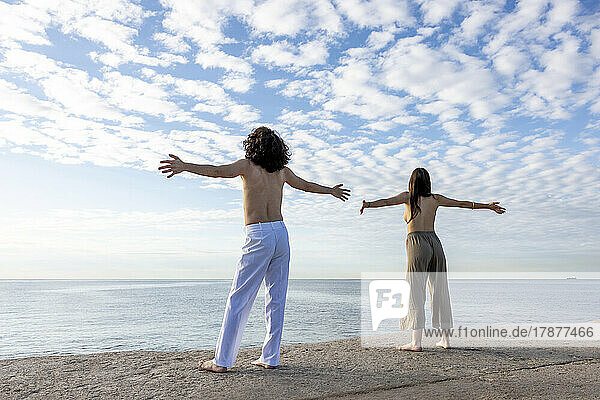 Carefree young couple standing topless with arms outstretched on pier