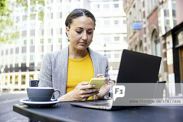 Businesswoman using smart phone sitting with laptop at sidewalk cafe