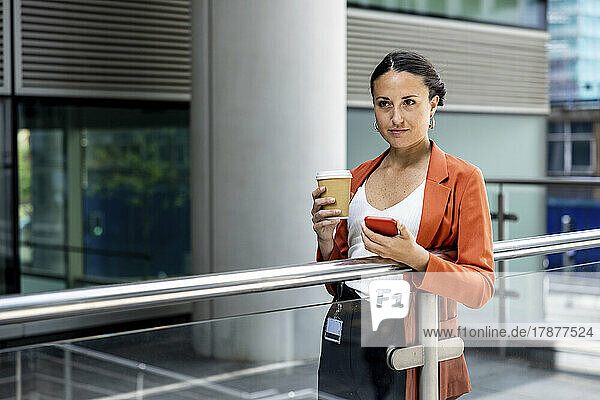 Businesswoman holding disposable cup and smart phone standing by railing
