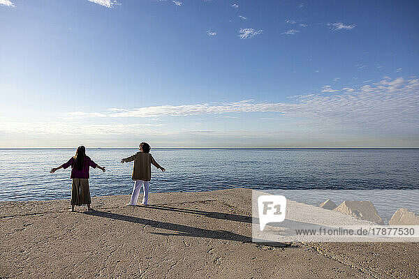 Couple standing on pier with arms outstretched looking at sea