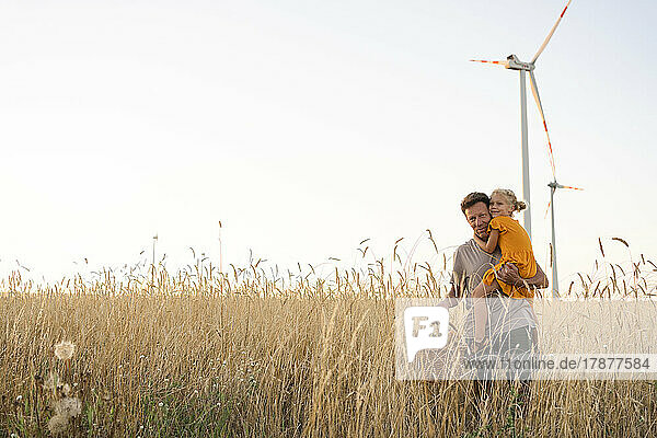 Father carrying daughter at wheat field with wind turbines