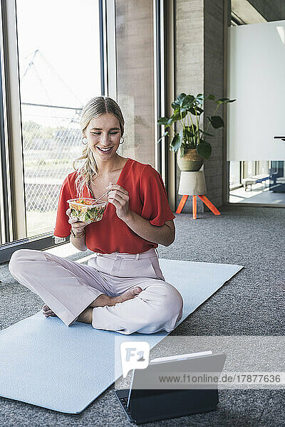 Smiling businesswoman eating salad watching tablet PC on exercise mat in office