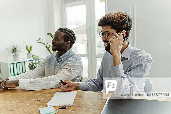 Happy businessman talking on mobile phone sitting by colleague at office