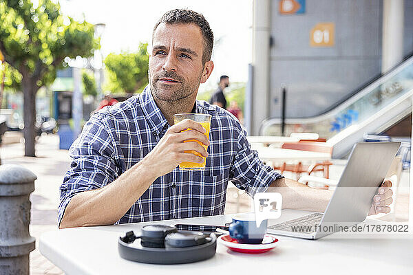 Mature freelancer sitting with laptop drinking orange juice at outdoor cafe