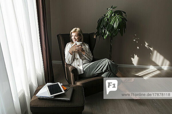 Happy mature woman having coffee sitting in armchair at home
