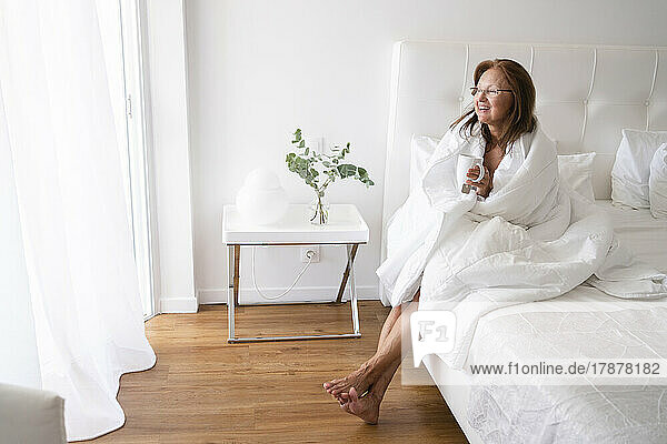 Smiling senior woman wrapped in blanket drinking tea sitting on bed