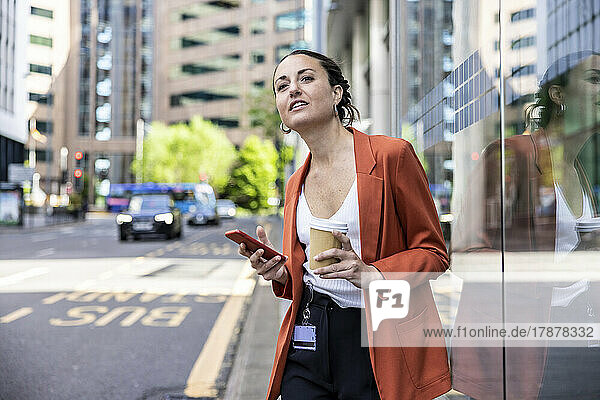 Businesswoman holding disposable cup and smart phone standing by glass wall