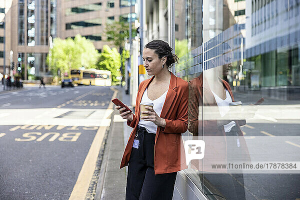 Businesswoman holding disposable cup using smart phone leaning on glass wall at sidewalk