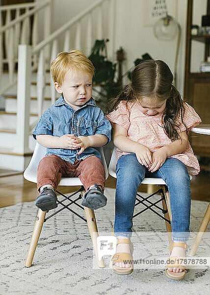 Pouting brother (12-17 months) and sister (2-3) sitting on chairs