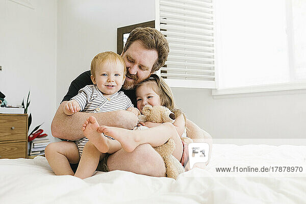 Father embracing son (12-17 months) and daughter (2-3) on bed