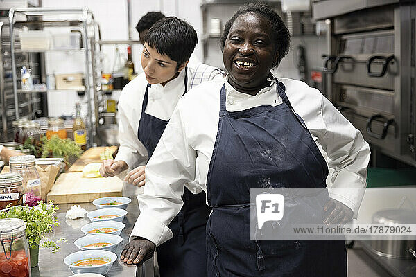 Porträt eines lächelnden Kochs mit einer weiblichen Kollegin  die im Hintergrund in einer Restaurantküche arbeitet