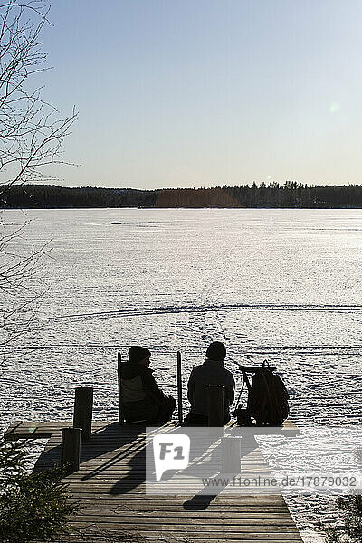 Rear view of boys sitting on jetty over frozen lake during sunny day
