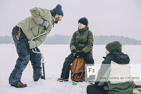 Happy mature man using ice auger while talking with son sitting on backpack at frozen lake