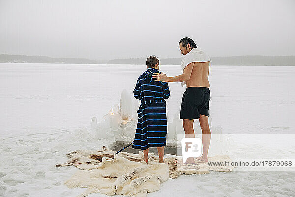 Rückansicht eines Mannes ohne Hemd  der sich mit seinem Sohn im Bademantel unterhält  während er auf einem Tierfell auf einem zugefrorenen See steht