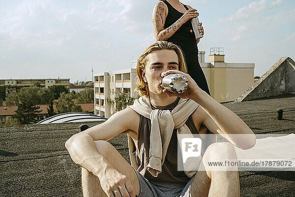 Junger Mann trinkt Wasser  während er vor einem Freund auf dem Dach sitzt