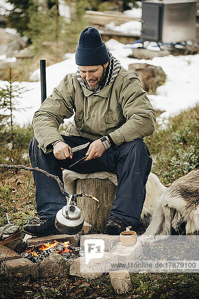 Lächelnder Mann in warmer Kleidung schaut auf Teekessel am Lagerfeuer
