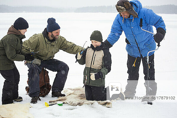 Happy mature men looking at boy fishing while kneeling on frozen lake during winter