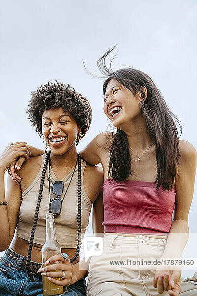 Cheerful young women laughing while sitting against sky