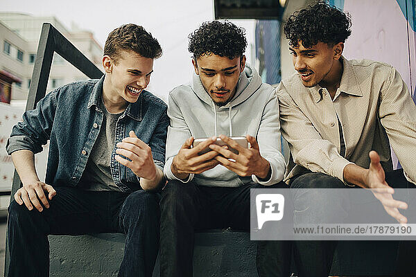 Junge Männer sehen einen Freund an  der ein Smartphone benutzt  während sie im Freien sitzen