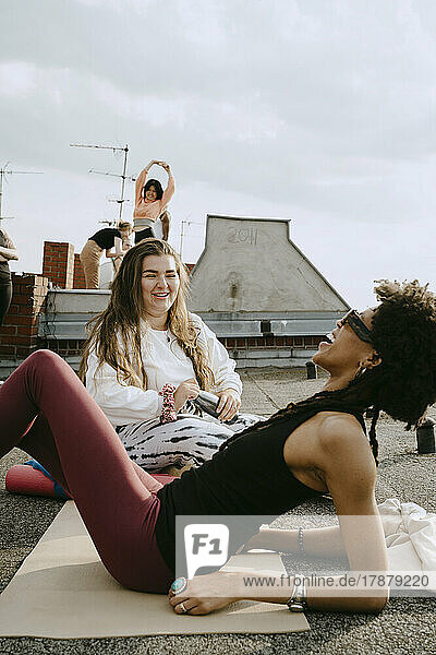 Lächelnde junge Frau im Gespräch mit einer fröhlichen Freundin auf einer Übungsmatte liegend