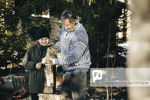 Lächelnder reifer Mann  der seinem Sohn beibringt  einen Baumstamm mit einer Axt im Wald zu fällen