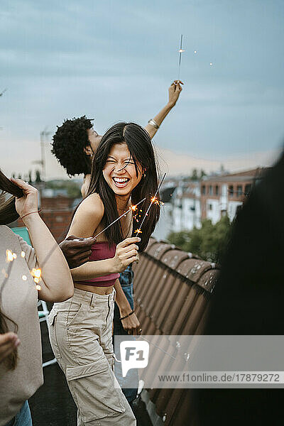 Cheerful young woman holding lit sparkler enjoying with friends on rooftop