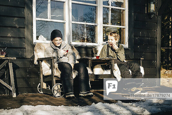 Lächelnde Freunde in voller Länge auf Stühlen sitzend auf einer Veranda im Winter