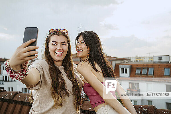 Glückliche Frauen strecken ihre Zunge heraus  während sie ein Selfie mit ihrem Smartphone auf dem Dach machen