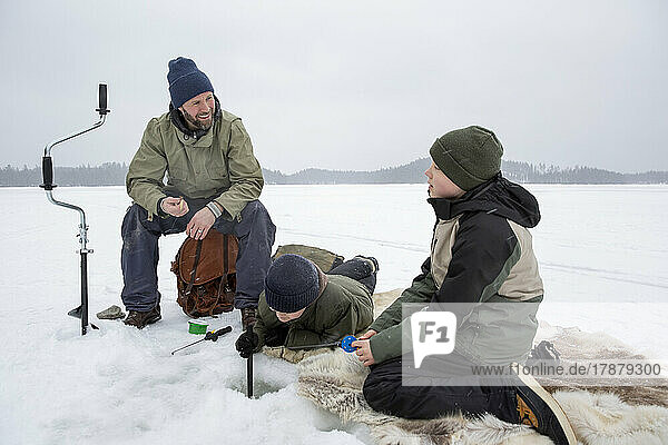 Lächelnder Mann im Gespräch mit einem Jungen  während sein Sohn auf einem zugefrorenen See Eisfischen geht