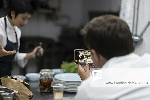 Männlicher Kollege filmt Koch bei der Zubereitung von Speisen in der Küche eines Restaurants