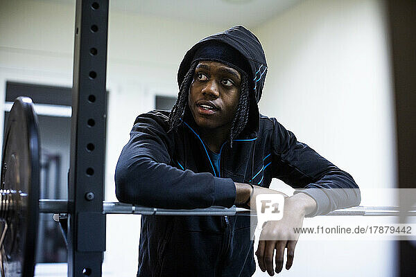 Männlicher Sportler schaut weg  während er sich im Fitnessstudio auf eine Hantel stützt