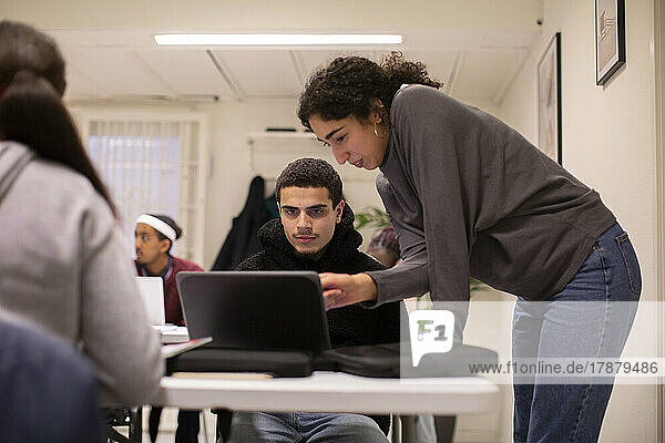 Lehrer lehnt sich an den Schreibtisch und hilft einem Schüler bei der Benutzung eines Laptops im Klassenzimmer