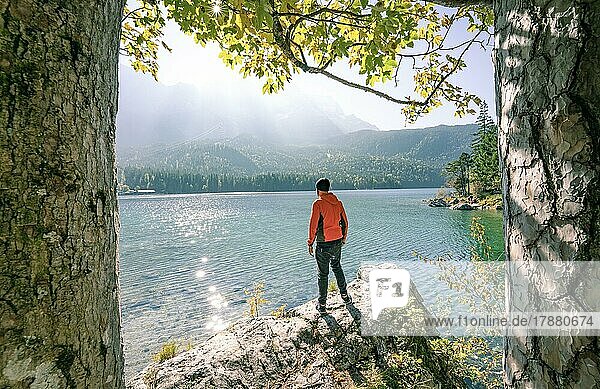 Mann in Orange steht auf einem Fels und schaut auf den See im ersten Sonnenlicht  Eibsee  Bayern  Deutschland  Europa