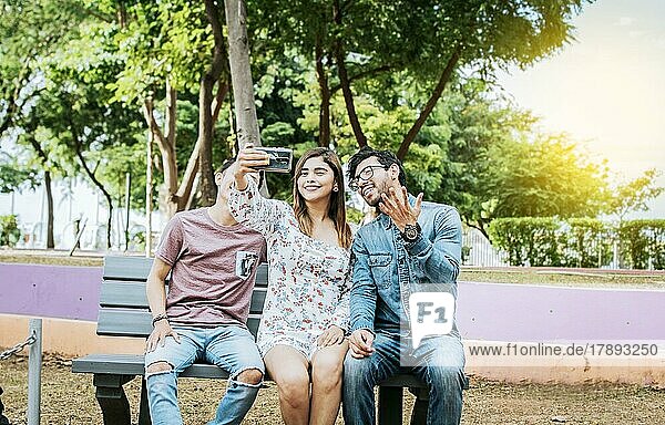 Three friends sitting on a bench taking a selfie  Three teenage friends sitting on a bench taking selfies. Gathering of three happy friends taking a selfie sitting on a bench