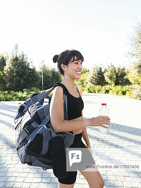 Happy young sportswoman with gym bag walking on footpath