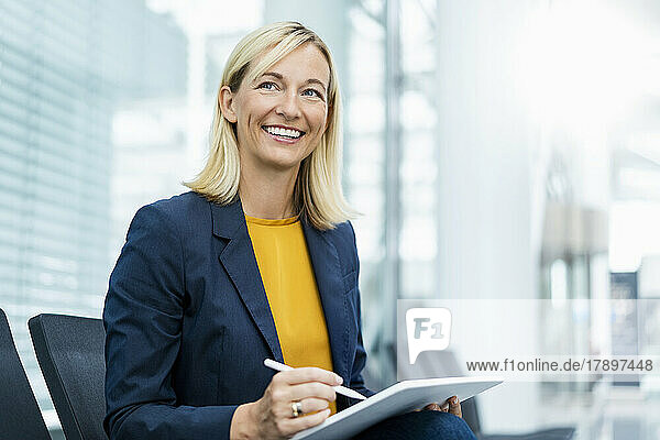 Smiling businesswoman with tablet PC sitting on chair in lobby