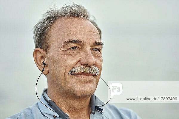 Thoughtful senior man with in-ear headphones listening music in front of sea