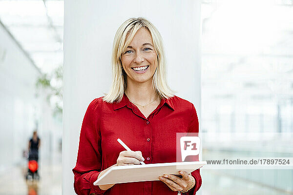 Smiling businesswoman with tablet PC and digitized pen standing in front of column