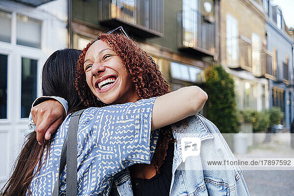 Happy woman with curly hair hugging friend in front of building