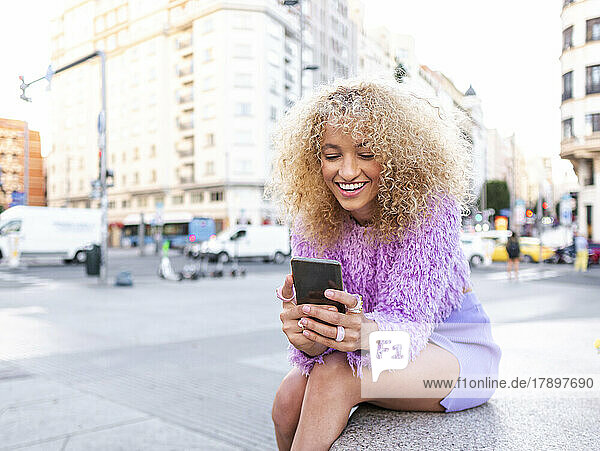 Lächelnde junge Frau  die ihr Mobiltelefon benutzt und auf einer Bank in der Stadt sitzt