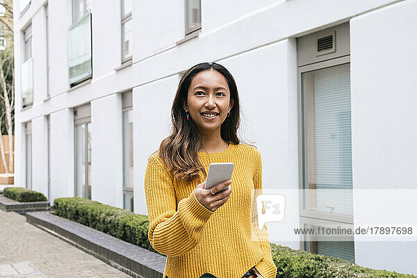 Happy young woman holding mobile phone by building