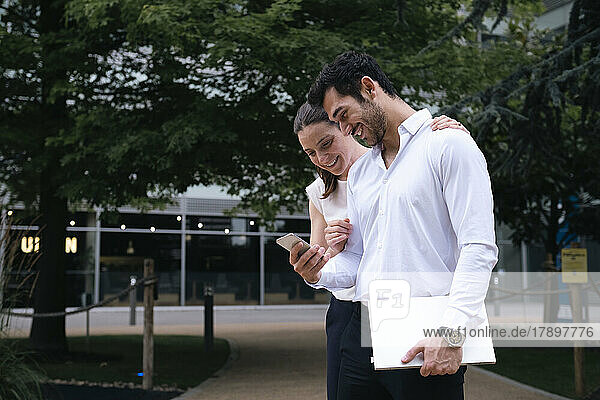 Smiling young businessman sharing mobile phone with businesswoman at office park