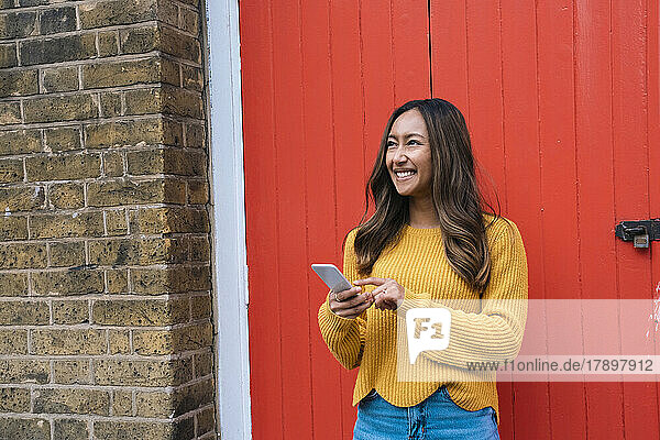 Glückliche junge Frau mit Mobiltelefon steht vor roter Tür