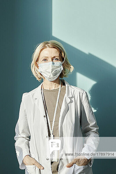 Mature doctor with protective face mask in front of wall