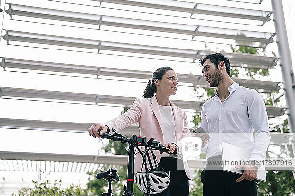 Lächelnde Geschäftsfrau mit Fahrrad im Gespräch mit Kollegen im Büropark