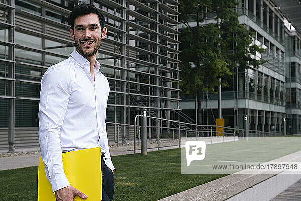Happy young businessman with file standing at office park